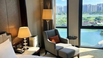 A hotel room with a bed, a chair, a lamp, a small table, and a large window offering a view of a cityscape and a body of water.