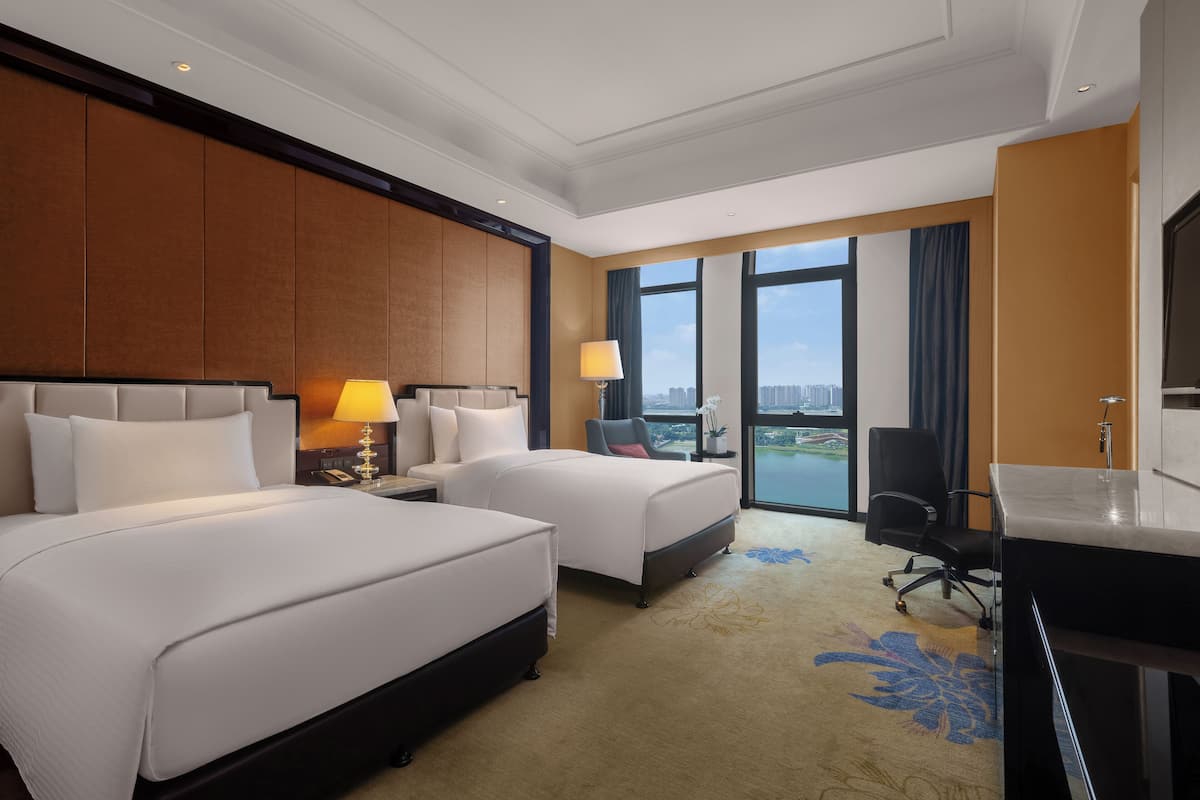 A hotel room with two beds, a desk, a chair, a large window with a view, and a carpet with a floral pattern.