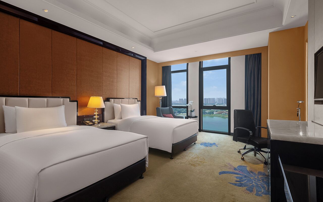 A hotel room with two beds, a desk, a chair, and a large window with a view.