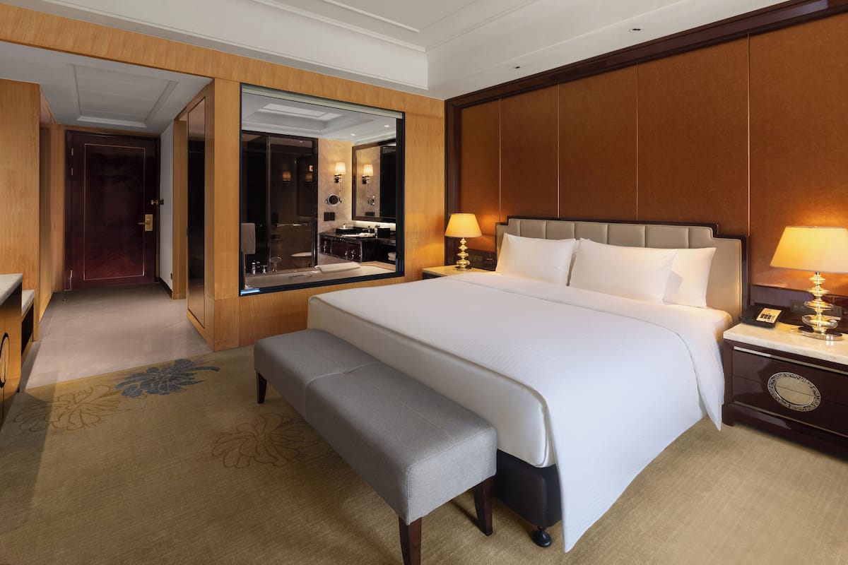 A hotel room with a large bed, a bench, a nightstand with a lamp, a mirror, and a wooden panel wall.