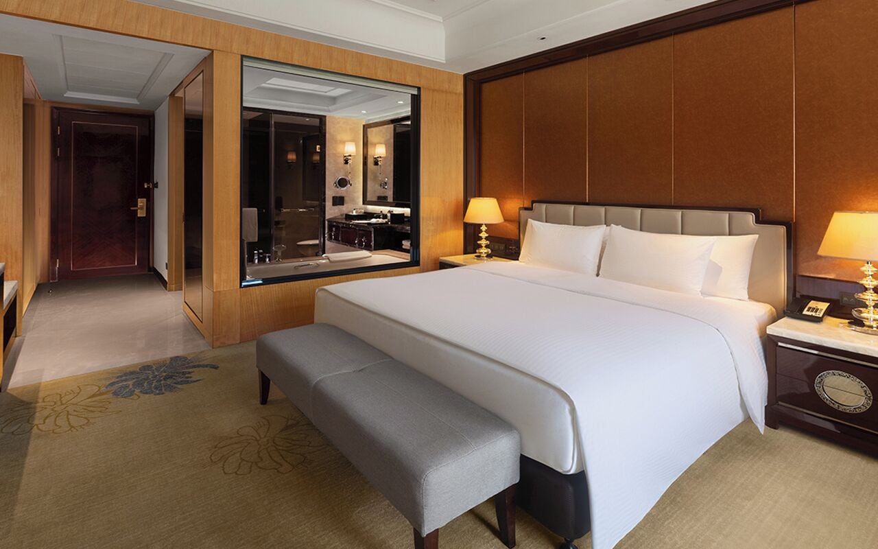 A hotel room with a large bed, a bench, a nightstand, a mirror, and a view of the bathroom.