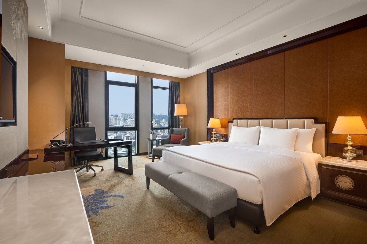 Habitación de hotel con una cama grande, un escritorio, una silla, un banco, una mesita de noche y vistas a la ciudad.