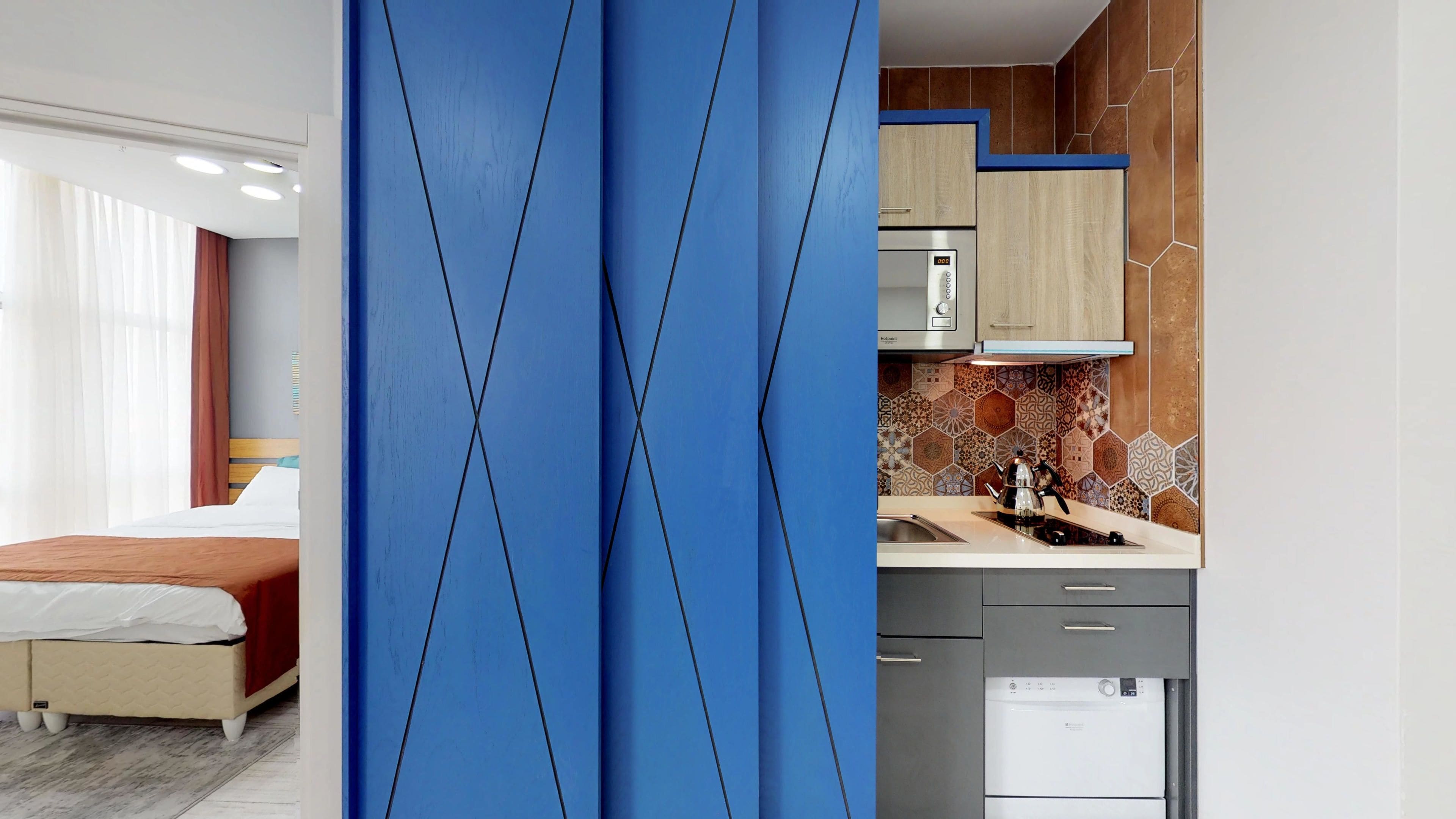 Une chambre d’hôtel moderne comprenant un lit, une cloison bleue, une kitchenette équipée d’un four à micro-ondes, d’une bouilloire et d’un lave-vaisselle.