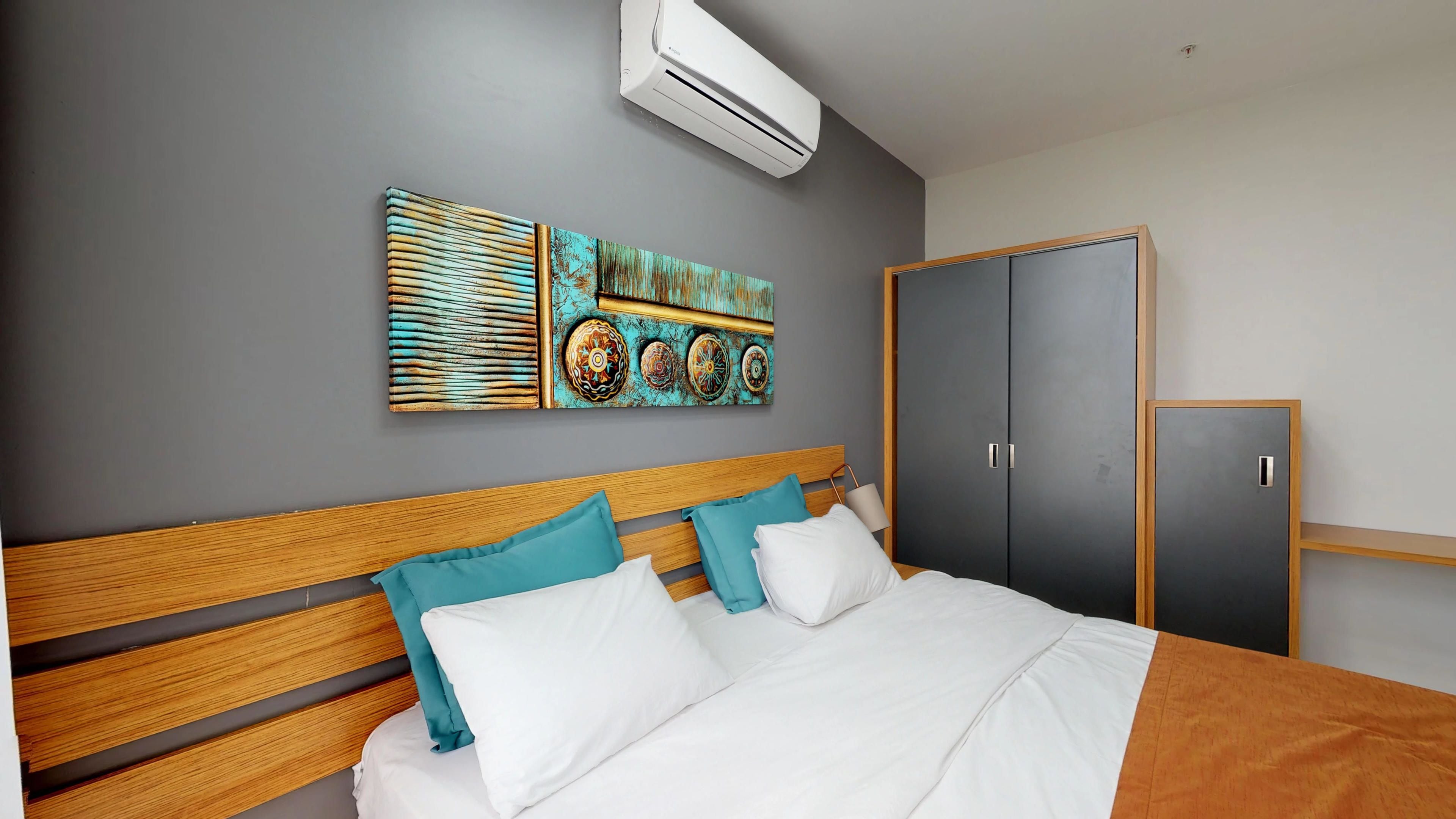 A hotel room with a wooden bed, teal pillows, a grey wardrobe, and a wall-mounted air conditioner.