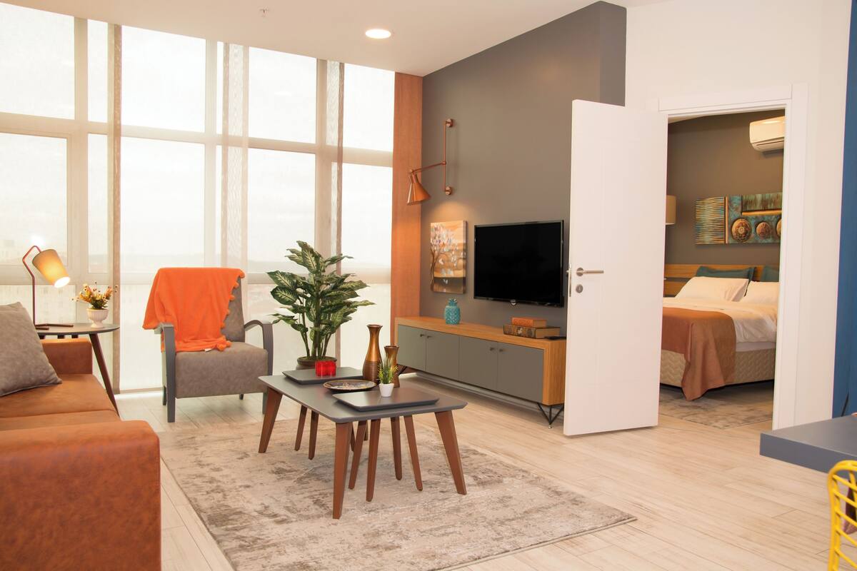 A modern living room with a sofa, a dining table, a TV, and a bed in the adjacent room.