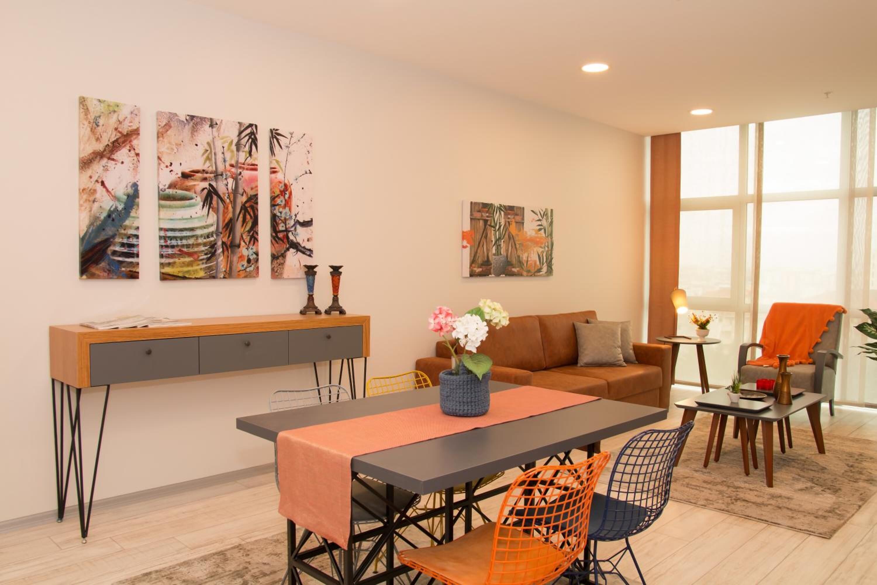 A modern living room with a dining area, featuring a sofa, a coffee table, and a dining table with chairs. There are abstract paintings on the wall and a window providing natural light.
