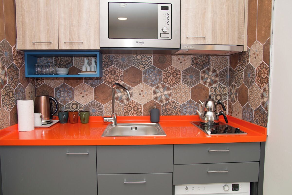 A kitchen with a microwave, sink, and orange countertop. There are cabinets and a blue shelf with glassware.