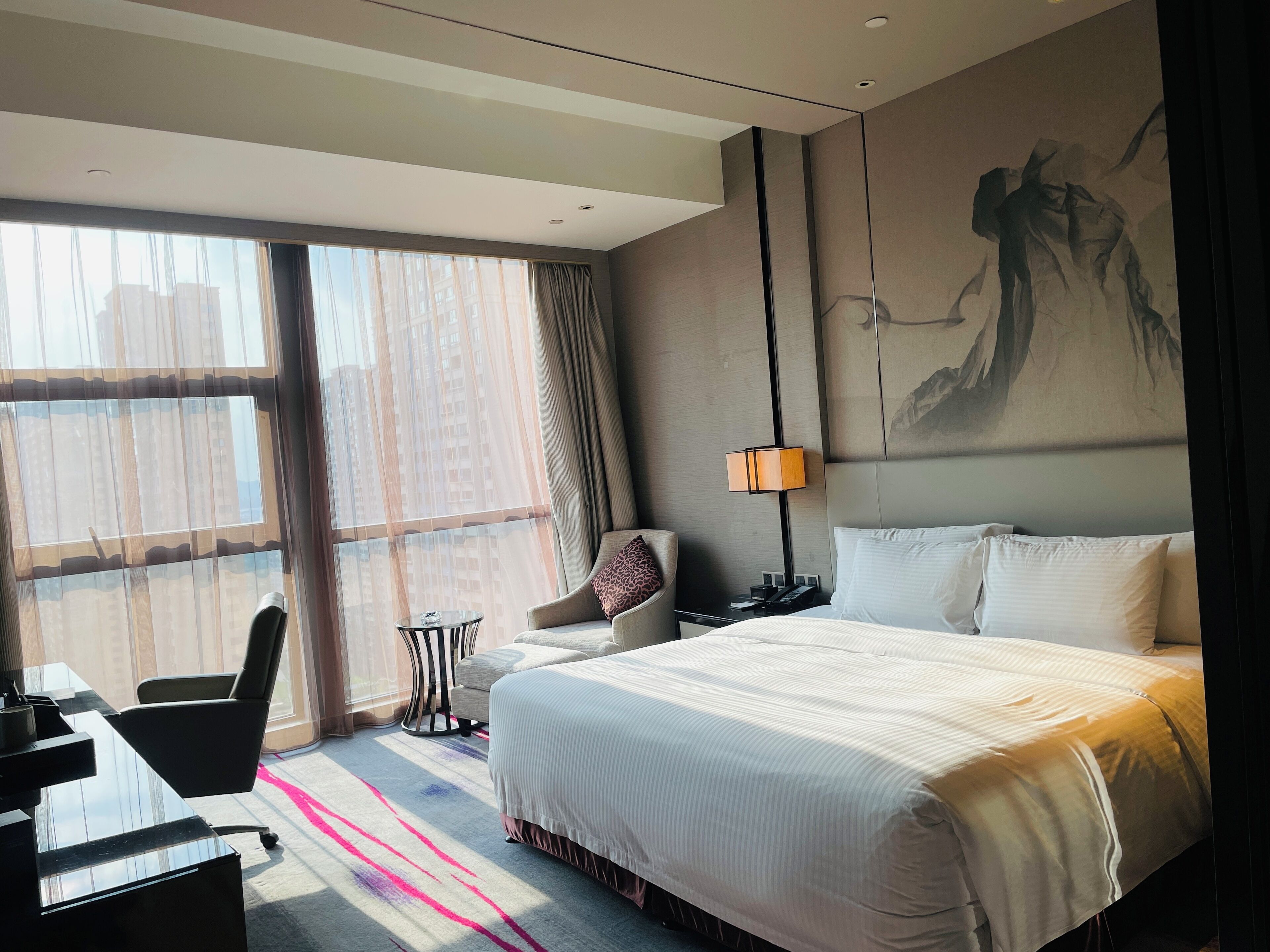 A hotel room with a large bed, a desk, a chair, a small table, and a large window with curtains.