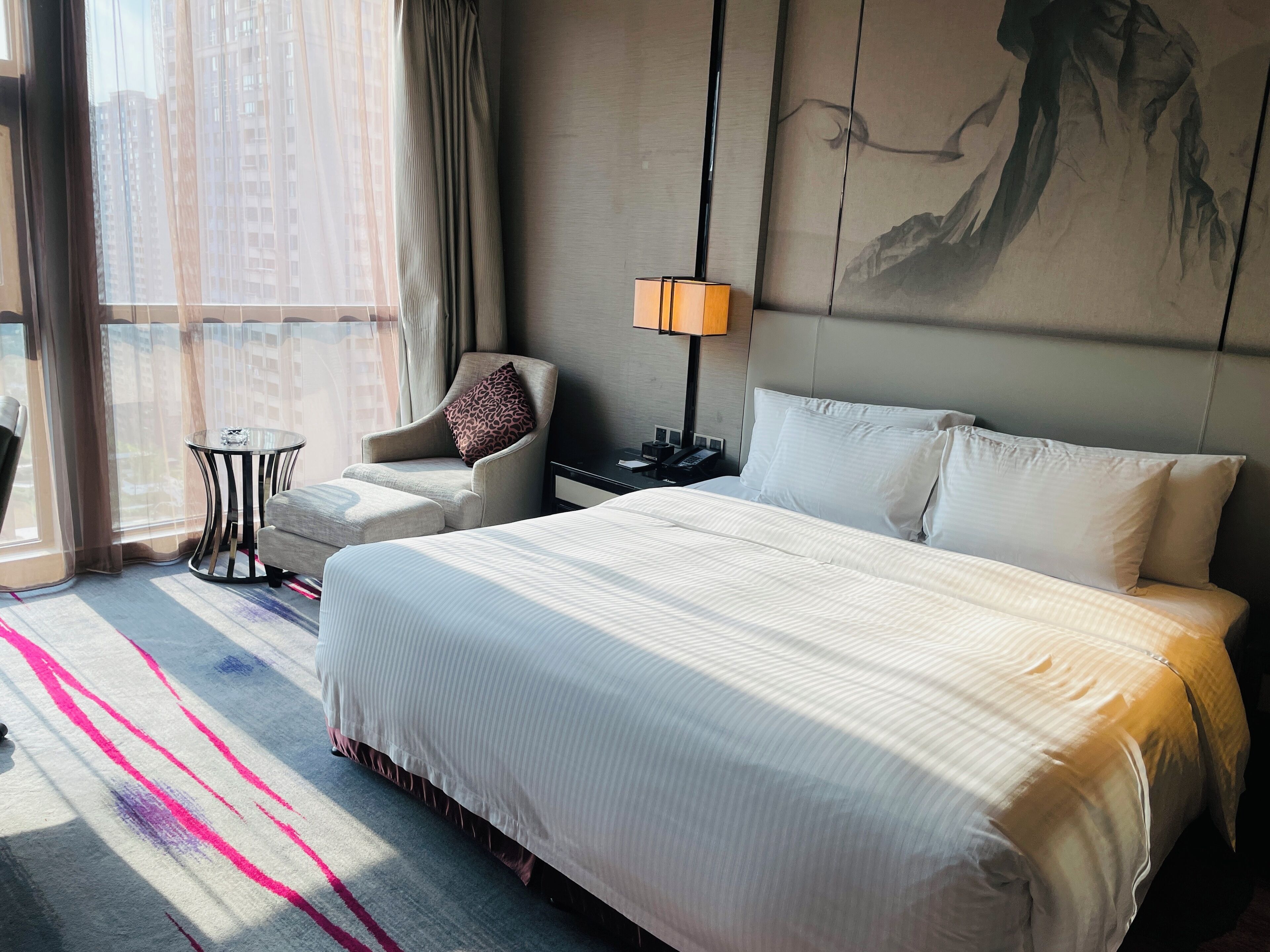 A hotel room with a large bed, a chair, a small table, a lamp, and a painting on the wall.