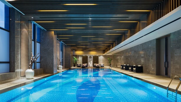 A modern indoor swimming pool with a sleek design, featuring a long, narrow pool with clear blue water and a reflection of the ceiling lights. - Wanda Realm Maanshan (Ma'anshan)