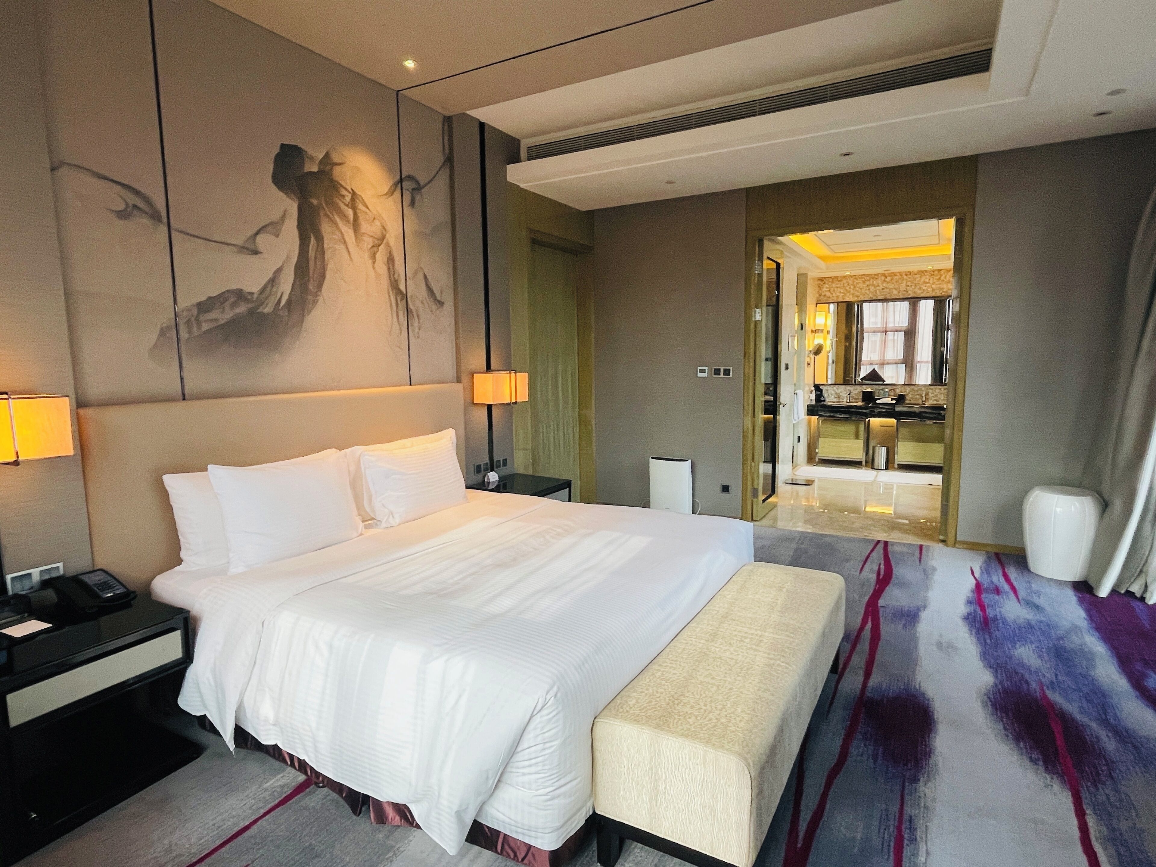 A hotel room with a large bed, bedside tables, a bench, a painting on the wall, and a view into the bathroom.