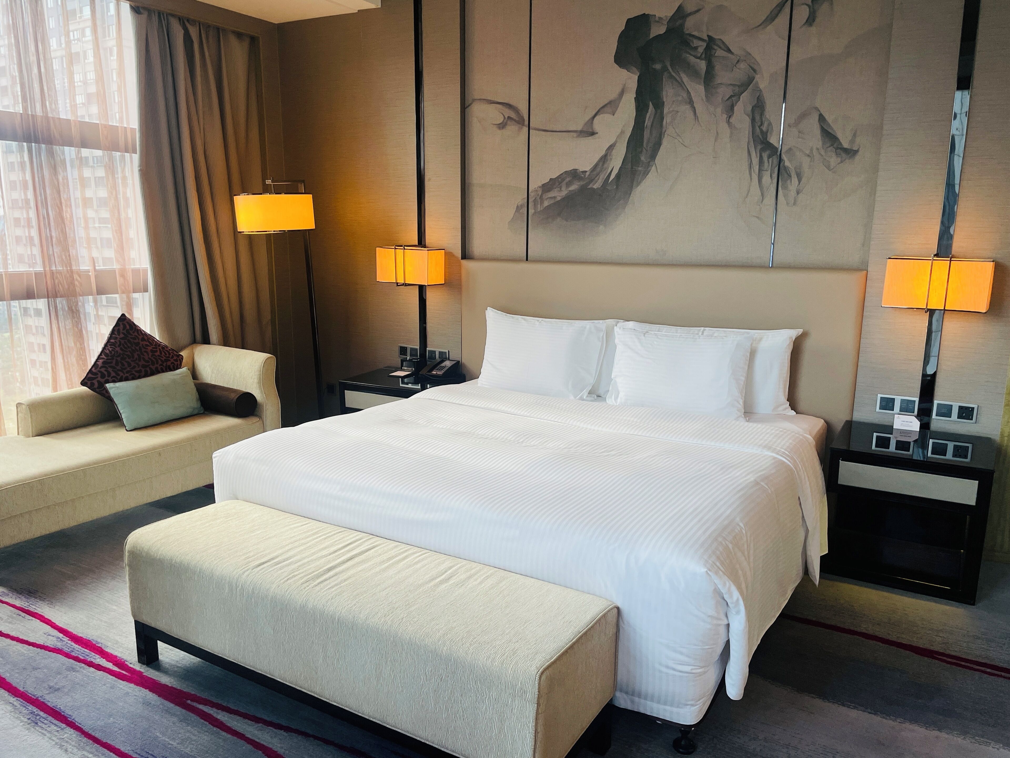 Une chambre d’hôtel avec un grand lit, deux tables de chevet avec des lampes, un canapé avec des coussins, un banc et un grand tableau abstrait accroché au mur.