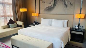 Une chambre d’hôtel avec un grand lit, deux tables de chevet avec des lampes, un canapé avec des coussins, un banc et un grand tableau abstrait accroché au mur.