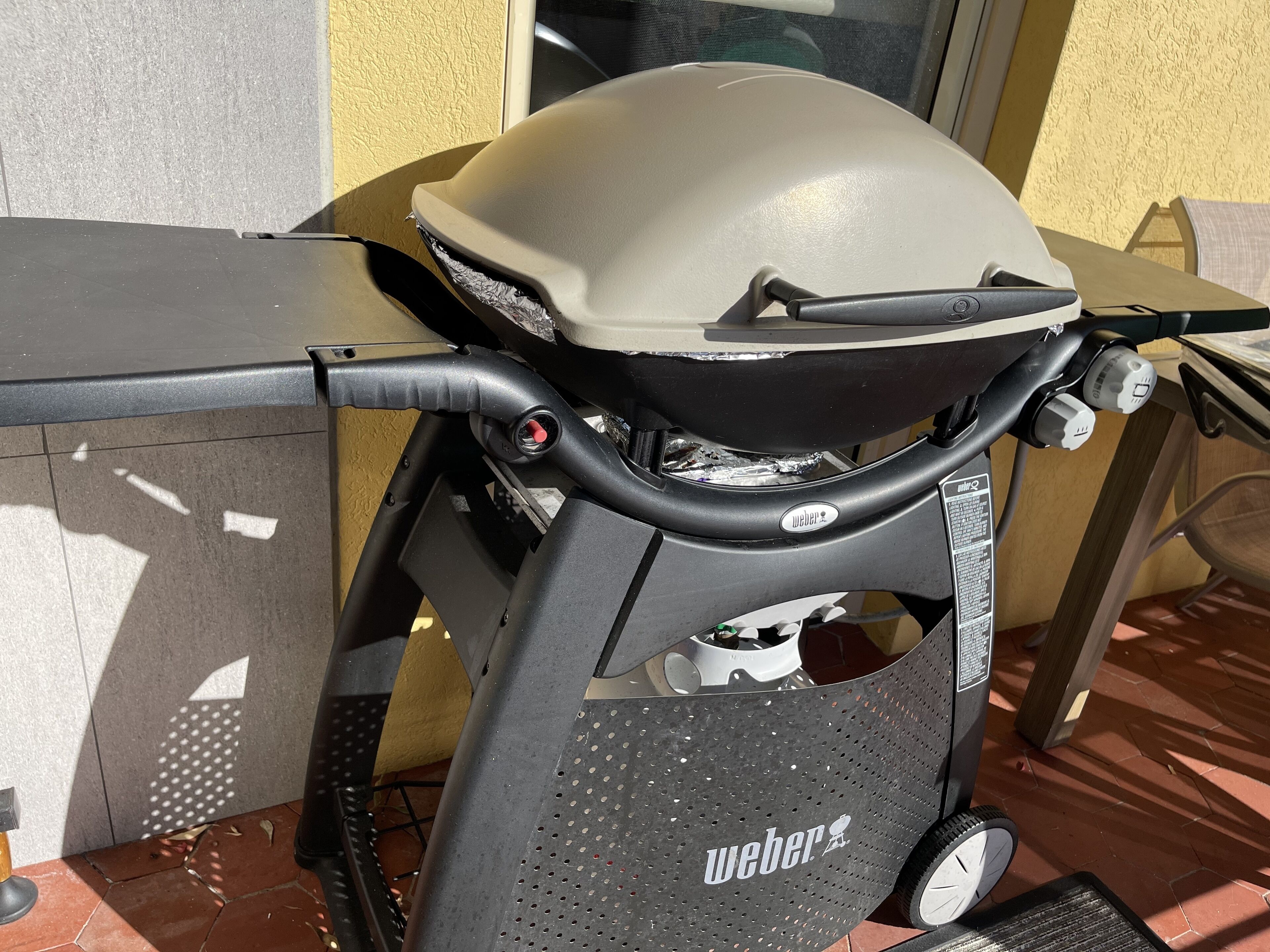 Una parrilla Weber portátil con un asa negra y un panel lateral perforado, ubicada en un patio con baldosas.
