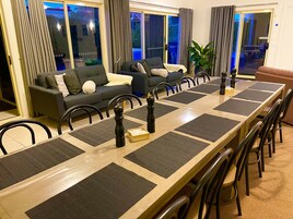 Un comedor con una larga mesa de madera, individuales negros y grises, y sillas negras. Se observa un sofá gris y una planta grande en maceta al fondo.