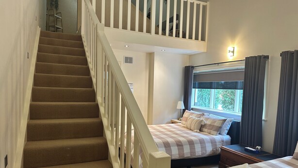 A bedroom with a checkered bedspread, a nightstand, a lamp, a window with curtains, and a staircase with a railing. - Austral Place 88 via Merri River (Dennington)