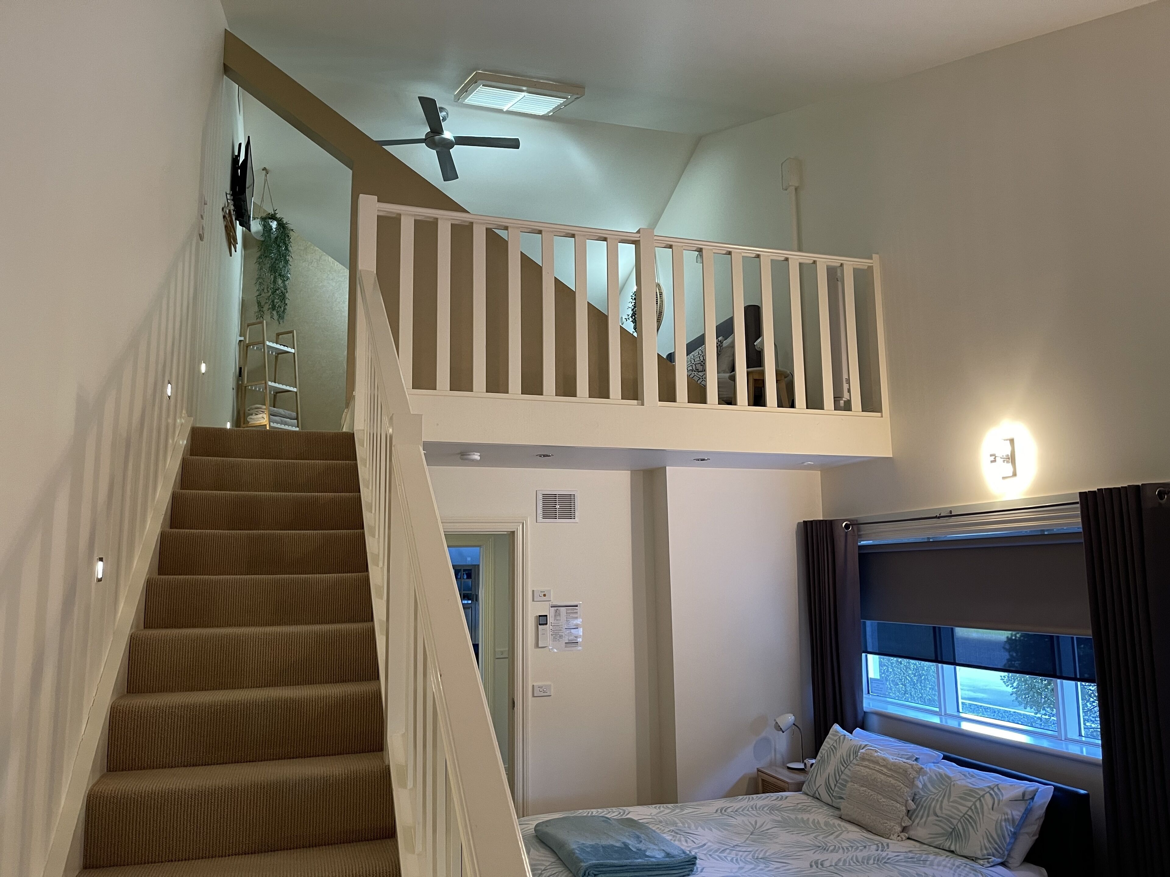 A bedroom with a bed, a window with curtains, a ceiling fan, and a staircase leading to an upper level.