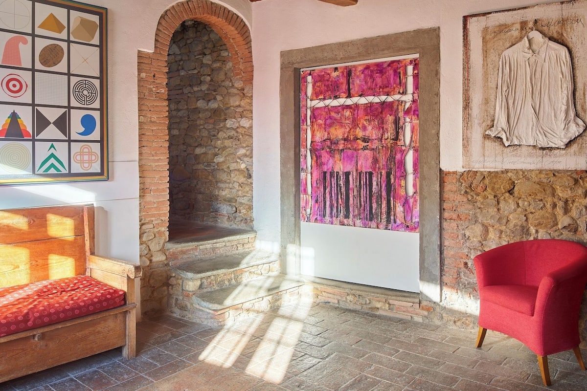 Una habitación con chimenea de piedra, un sillón rojo, un banco de madera y un cuadro abstracto de colores.