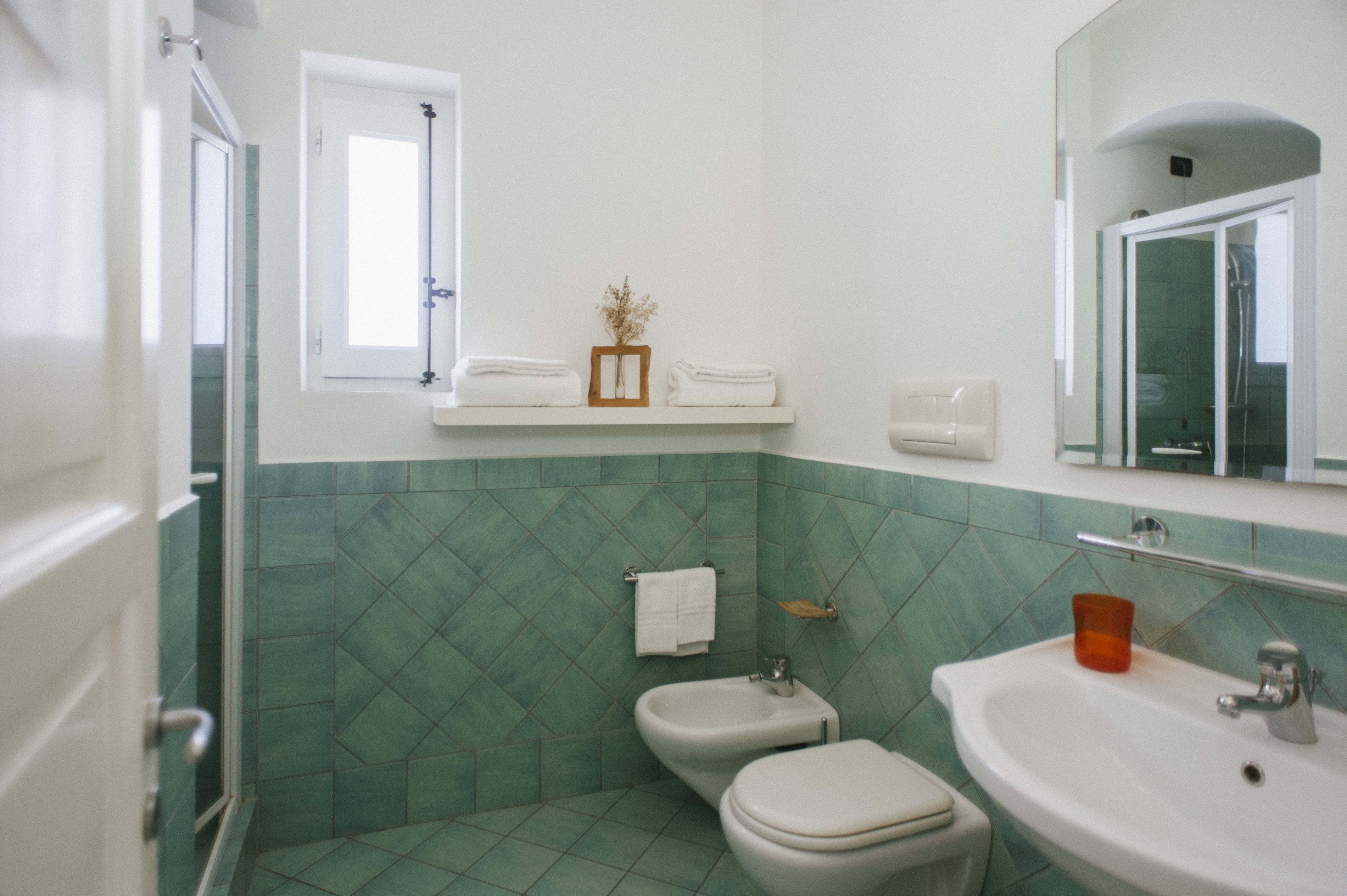 Un baño con pared de azulejos verdes, un lavabo blanco, un bidé y un inodoro. Hay una ventana sobre el lavabo y un espejo sobre el inodoro.
