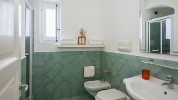 Un baño con pared de azulejos verdes, un lavabo blanco, un bidé y un inodoro. Hay una ventana sobre el lavabo y un espejo sobre el inodoro.