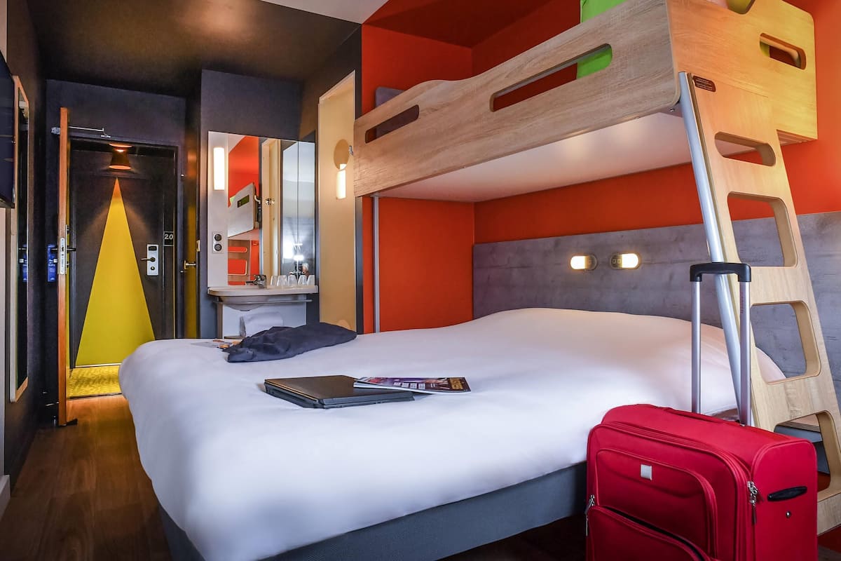 Une chambre d’hôtel avec un lit superposé, un bureau avec un miroir, une valise et un ordinateur portable posé sur le lit.