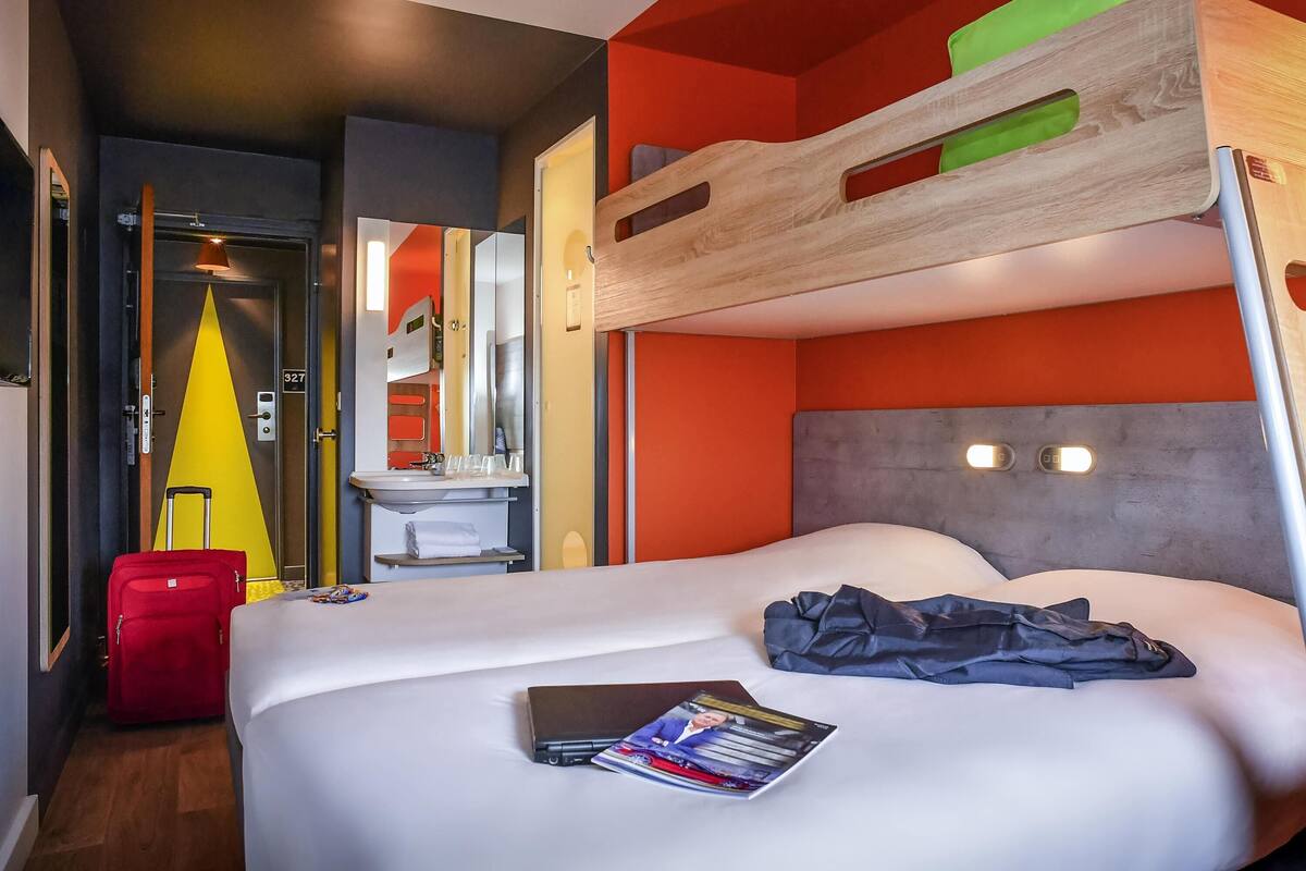 Une chambre d’hôtel avec un lit superposé, un lit simple, une salle de bain avec un lavabo et une valise.