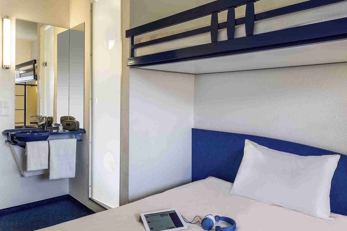 Une chambre d’hôtel avec un lit superposé, une table de chevet avec une tablette et des écouteurs, et une salle de bain avec un lavabo et un porte-serviettes.