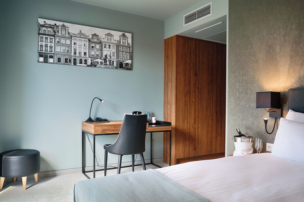 A hotel room with a bed, a desk with a chair, a wooden wardrobe, a wall-mounted lamp, and a painting of buildings.