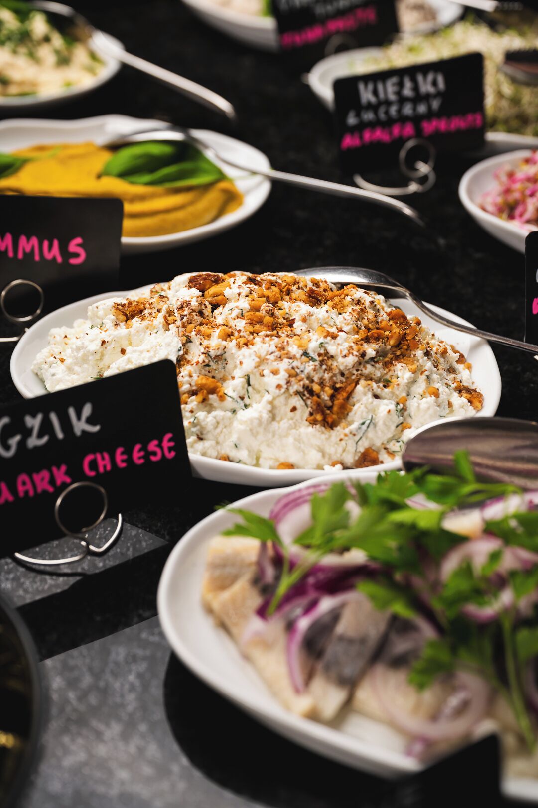 Una mesa con varios platos, incluyendo un plato con la etiqueta 'KIEK ALFA MESA' y otro con la etiqueta 'GZIK BLACK CHEESE'.
