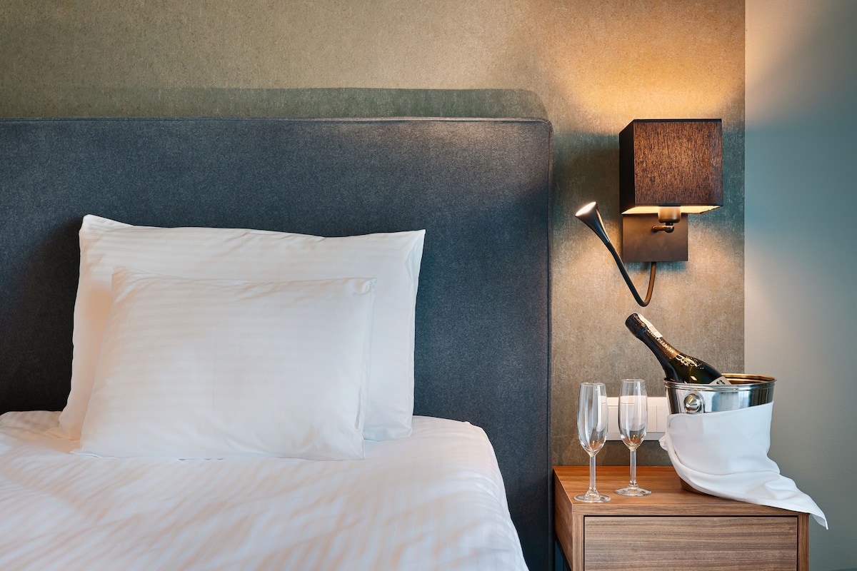 Una cama bien hecha con ropa de cama blanca, una mesita de noche con una botella de champán, dos copas y un pequeño tocador de madera.