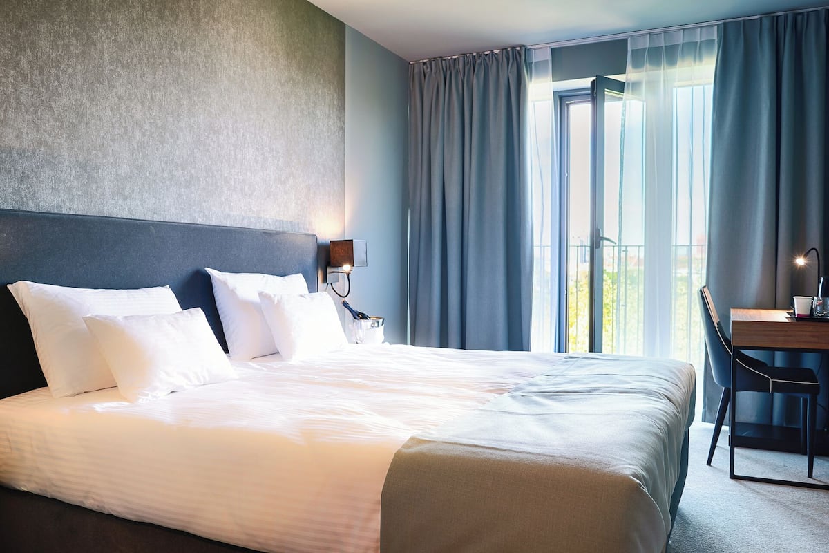 Una habitación de hotel con cama, escritorio, silla, mesita de noche y ventana con cortinas.