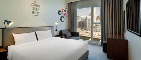 A hotel room with a large bed, a desk, a chair, and a view of the cityscape through the window.
