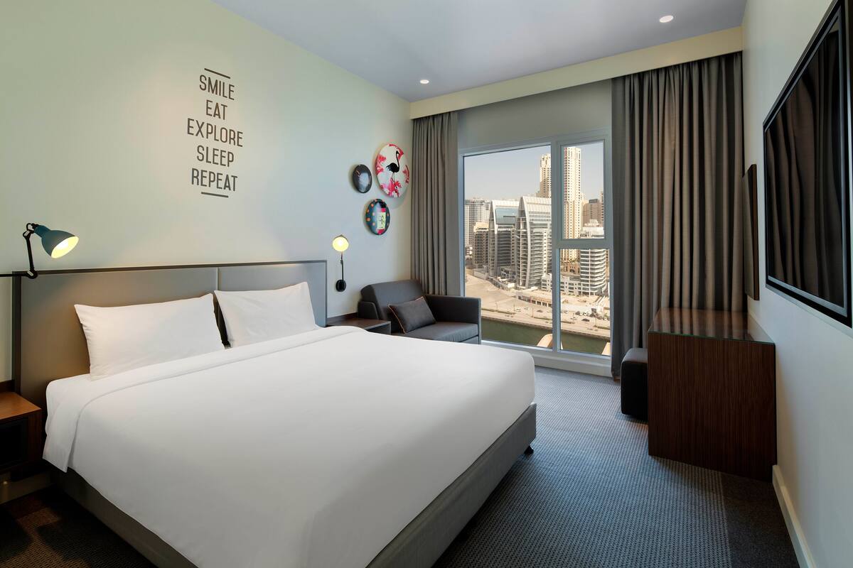 A hotel room with a large bed, a desk, a chair, and a view of the cityscape through the window.