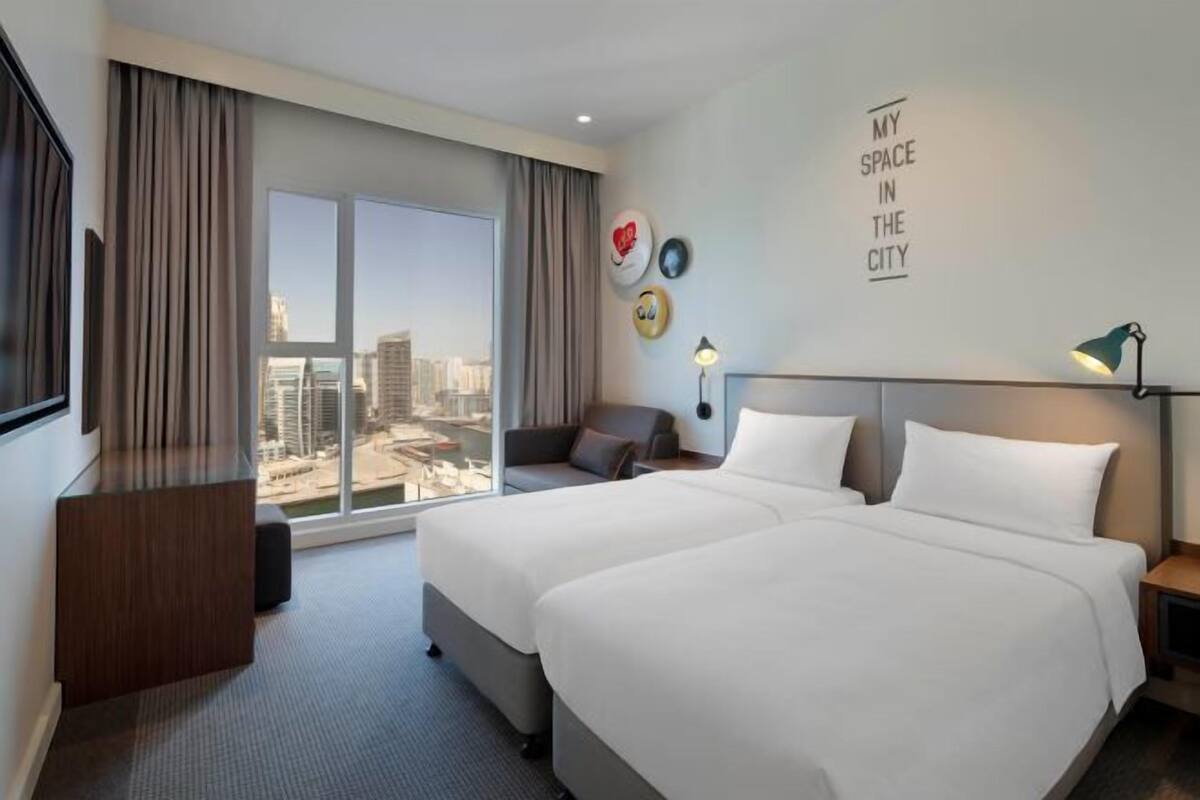 A hotel room with a large bed, a desk, a chair, a TV, a window with a city view, and a wall with a motivational quote.