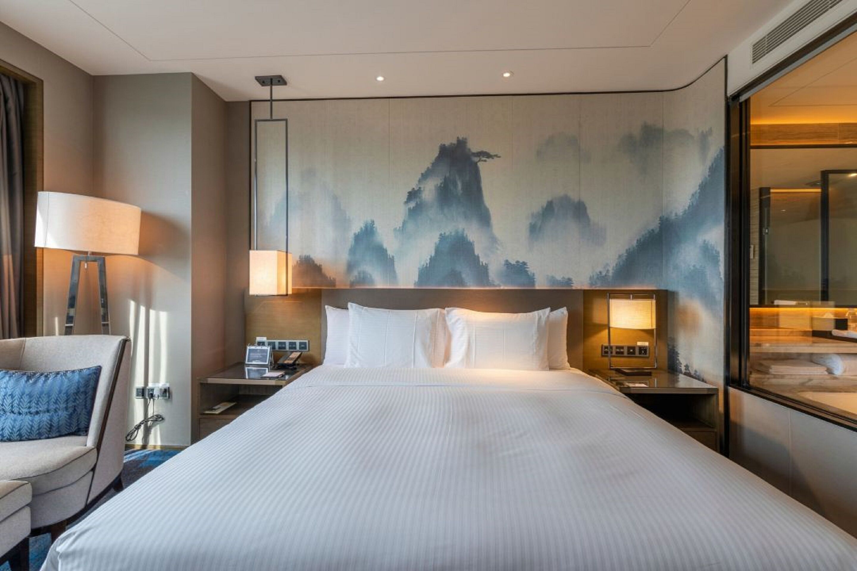 A hotel room with a large bed, a desk with a lamp, and a wall with a mountain painting.