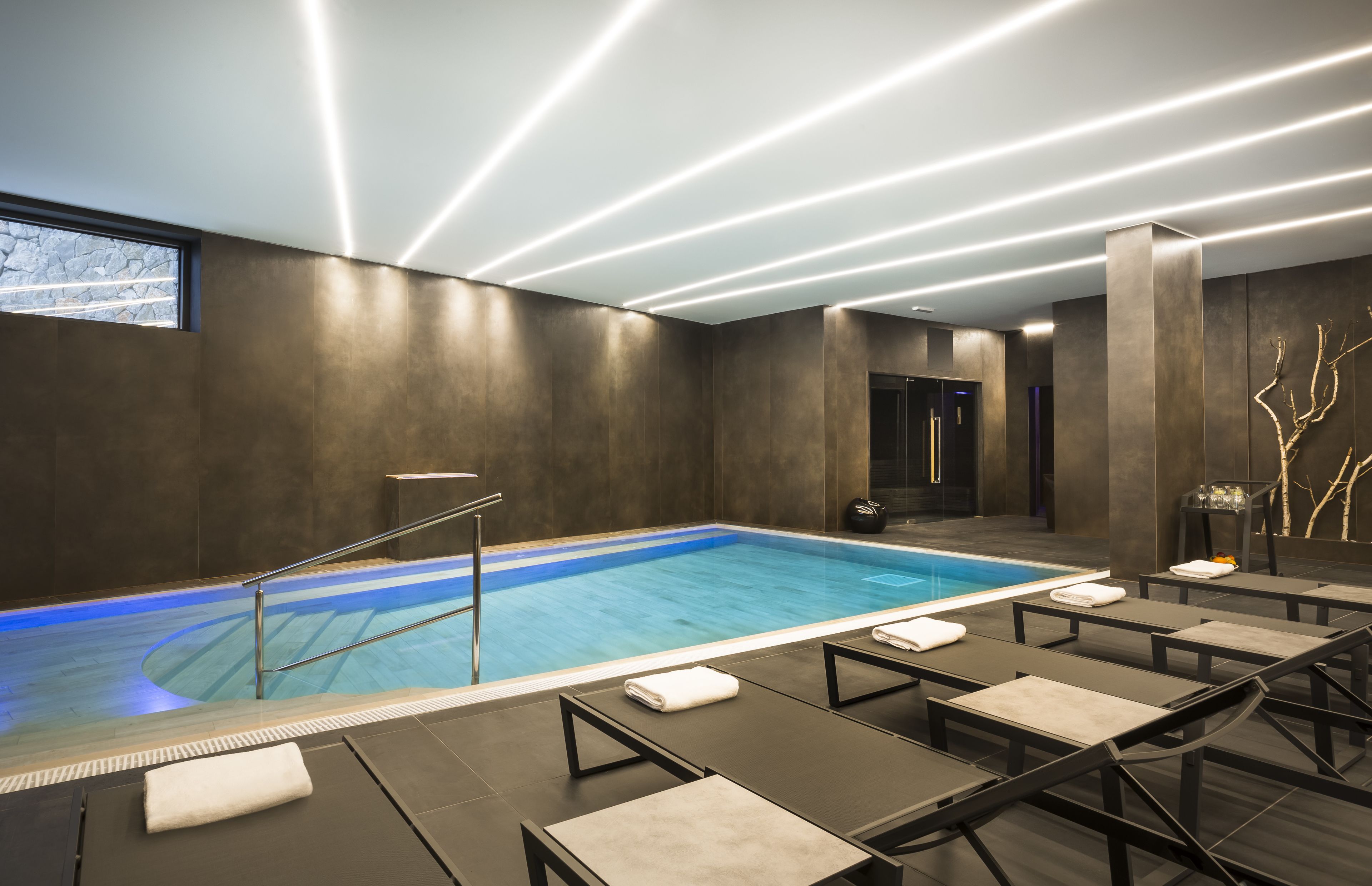 A modern indoor swimming pool with a clear blue water, surrounded by lounge chairs with towels, and a contemporary ceiling design with linear lighting.