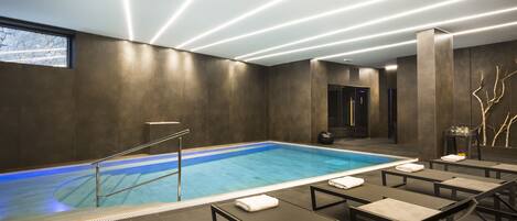 A modern indoor swimming pool with a clear blue water, surrounded by lounge chairs with towels, and a contemporary ceiling design with linear lighting.