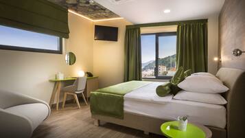 A hotel room with a bed, a desk, a chair, a small table, a TV, and a window with curtains.