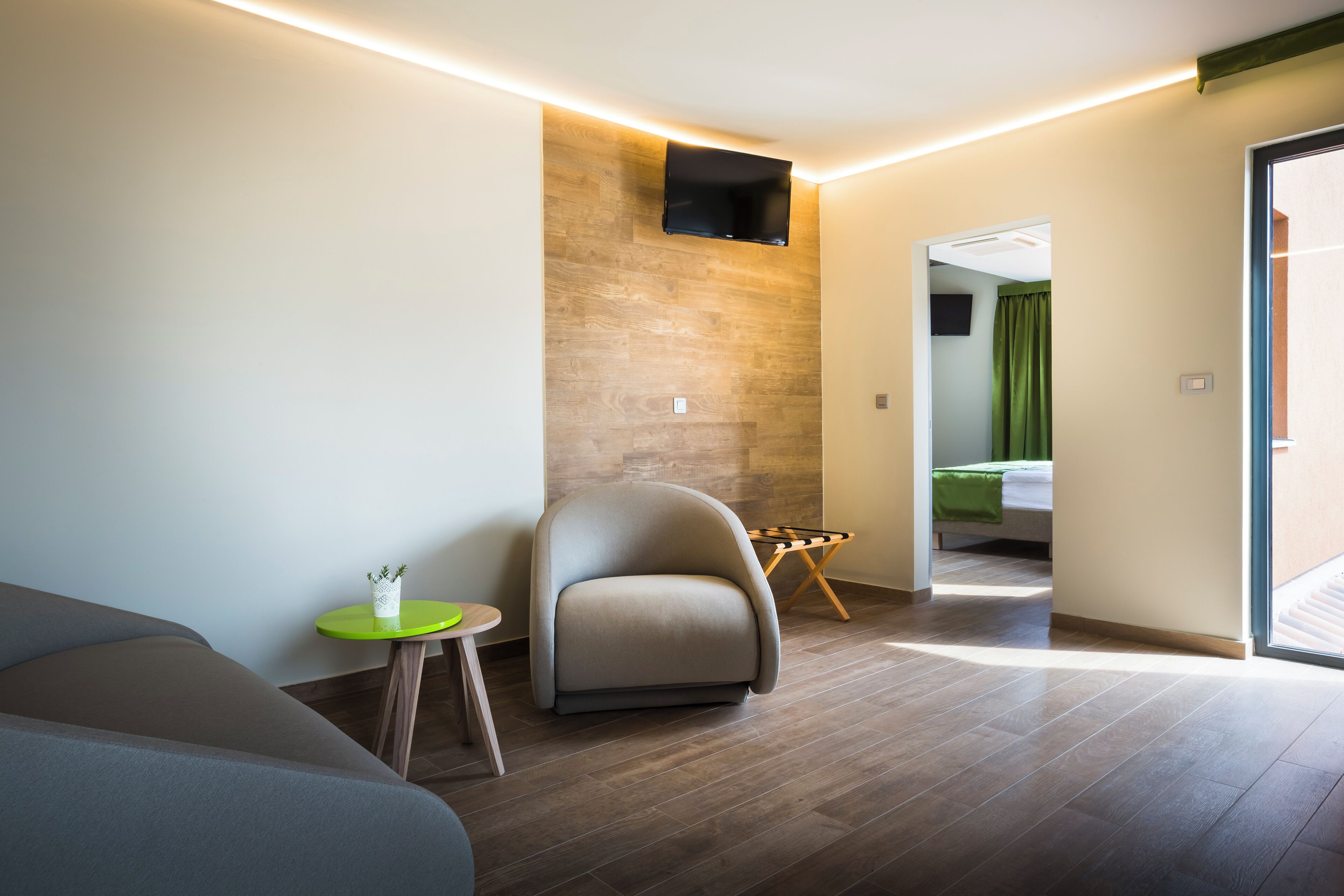 Et moderne hotelværelse med en sofa, et lille rundt bord med en plante, en stol, et fladskærms-tv ophængt på en trævæg og en seng med grønne gardiner.