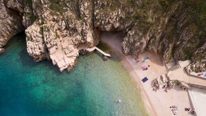 Vista aèria d'una zona costanera amb aigua turquesa cristal·lina, penya-segats rocosos i una platja sorrenca amb gent i cadires de bany.