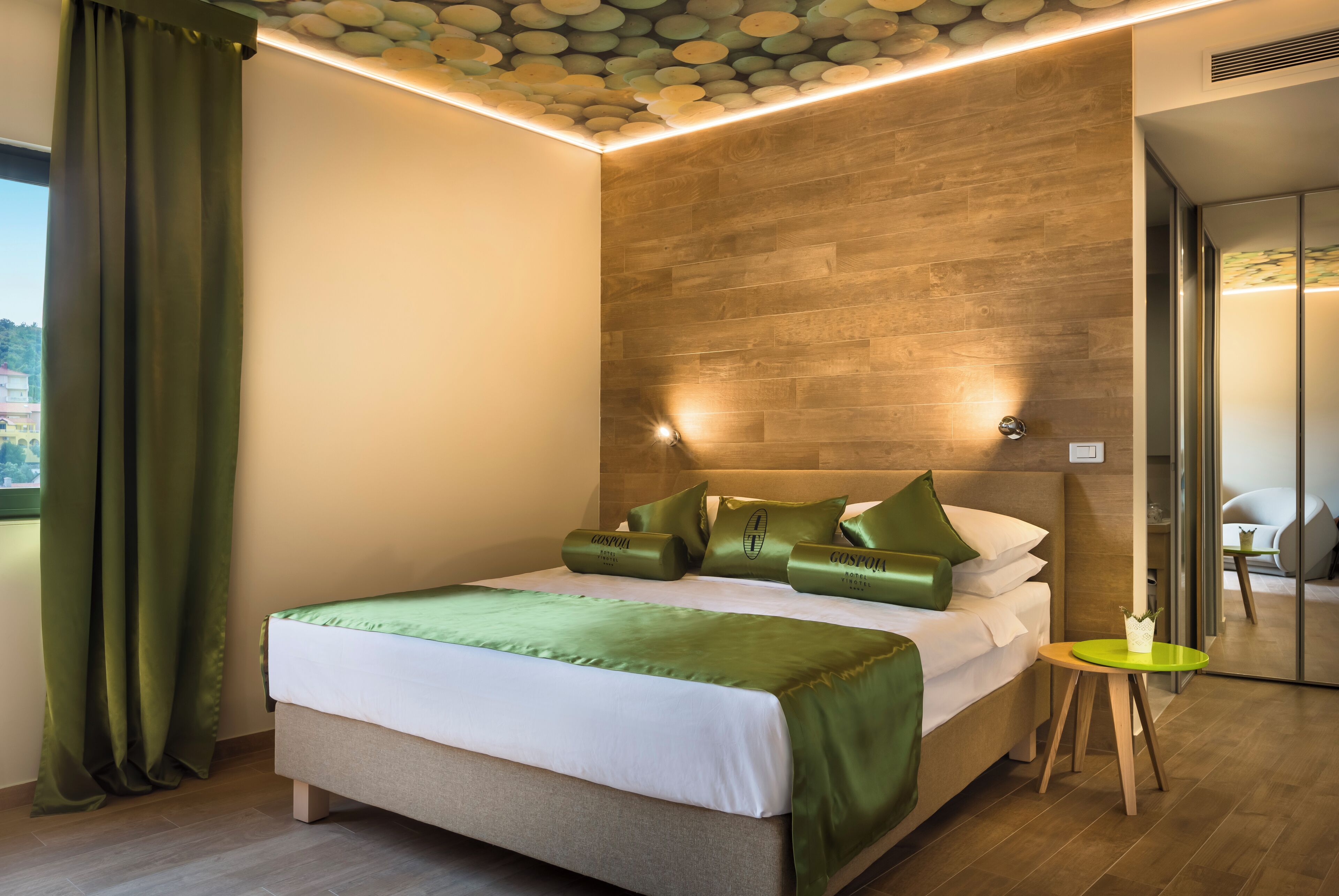 A modern hotel room with a wooden headboard, a neatly made bed with green bedding, a small round table, and a view of the outdoors through a window.