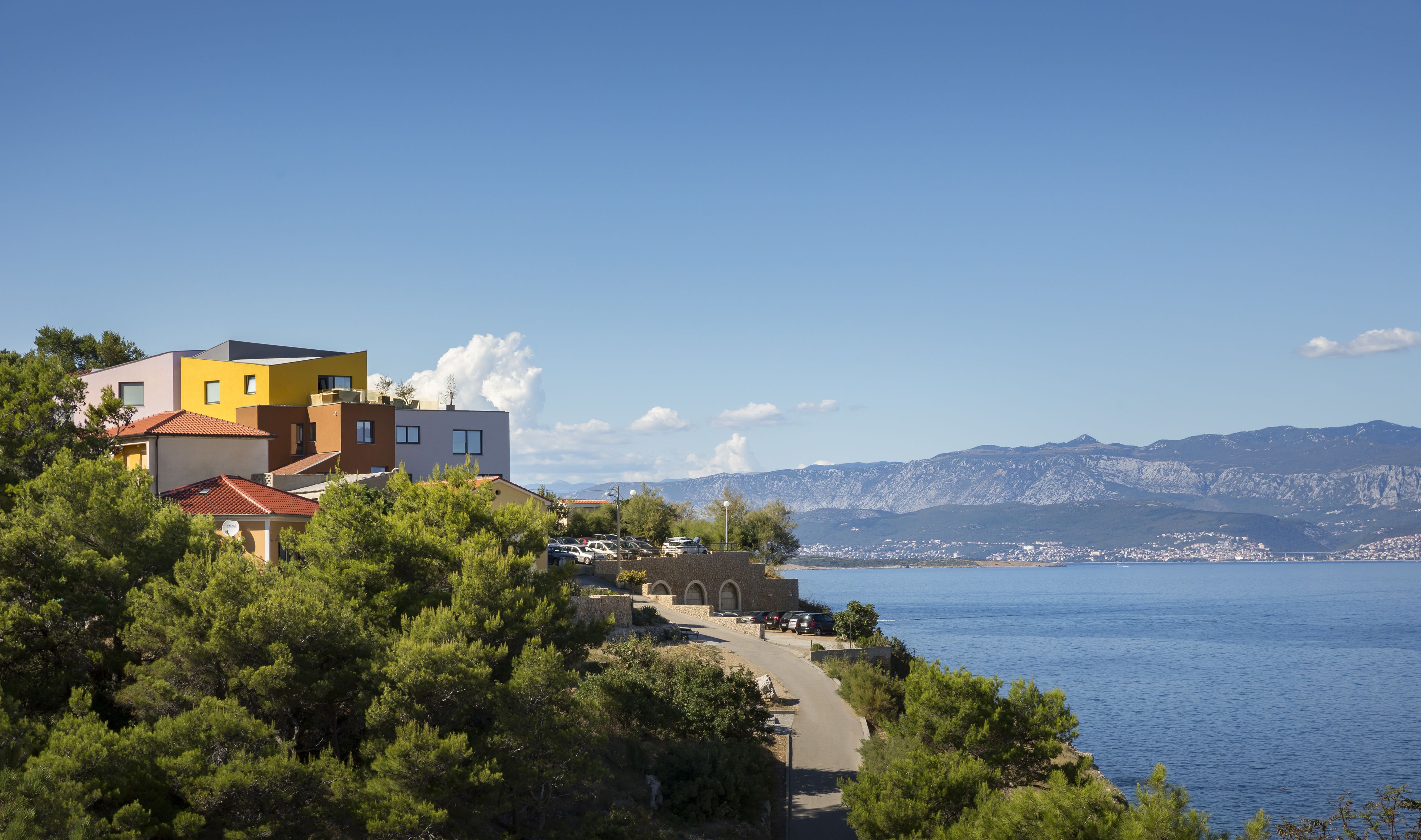 A coastal town with modern buildings, a clear blue sea, and mountains in the background.