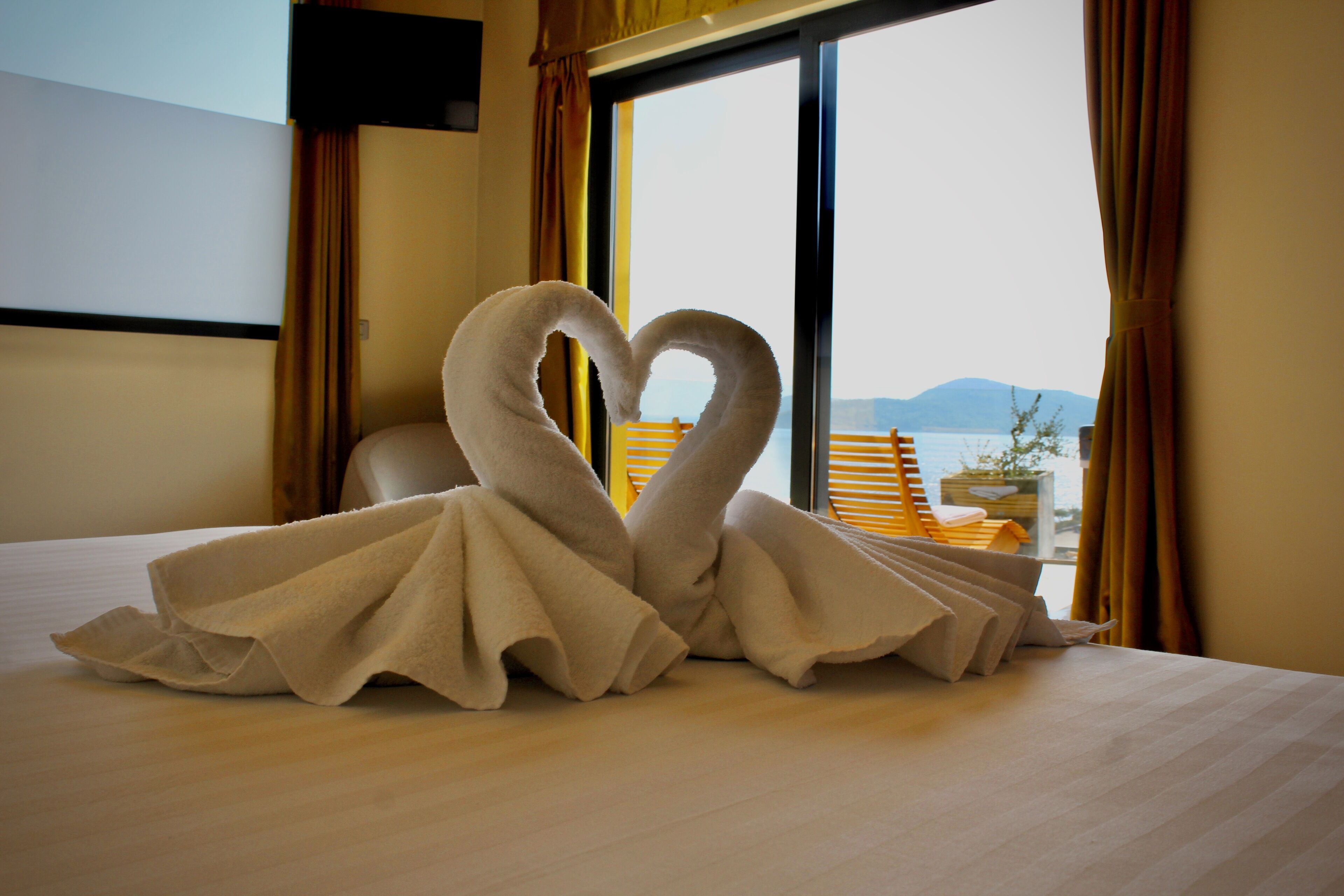 A bed with a heart-shaped towel arrangement and a view of a balcony with a wooden railing and a plant.