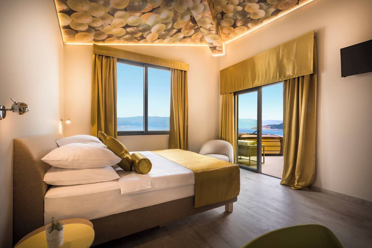 Habitación de hotel moderna con una cama grande, vista al exterior y un balcón con cortinas amarillas.