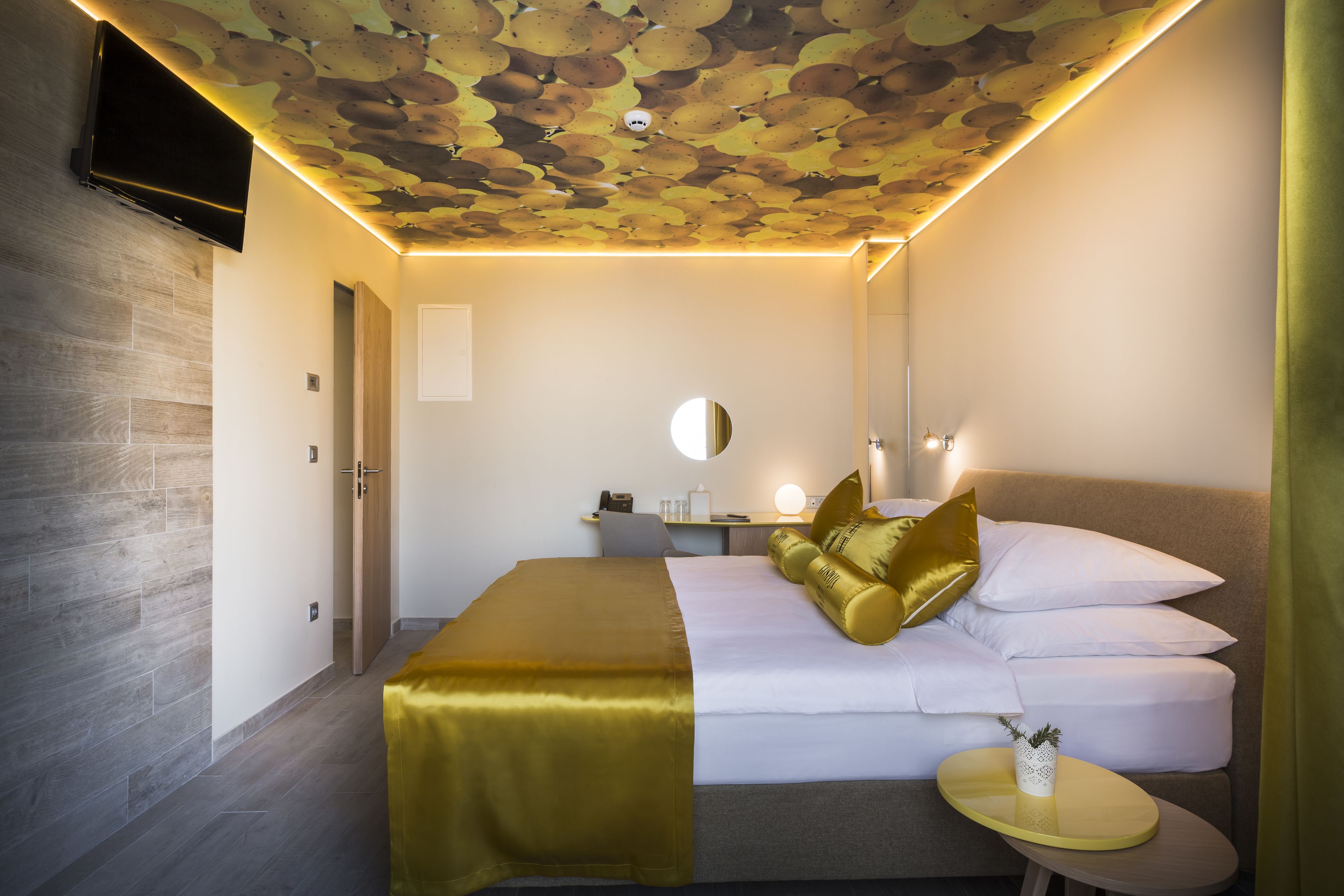 Habitación de hotel con diseño moderno, que presenta una cama con detalles dorados, un espejo redondo, una mesita de noche y un techo con un original patrón de hojas.