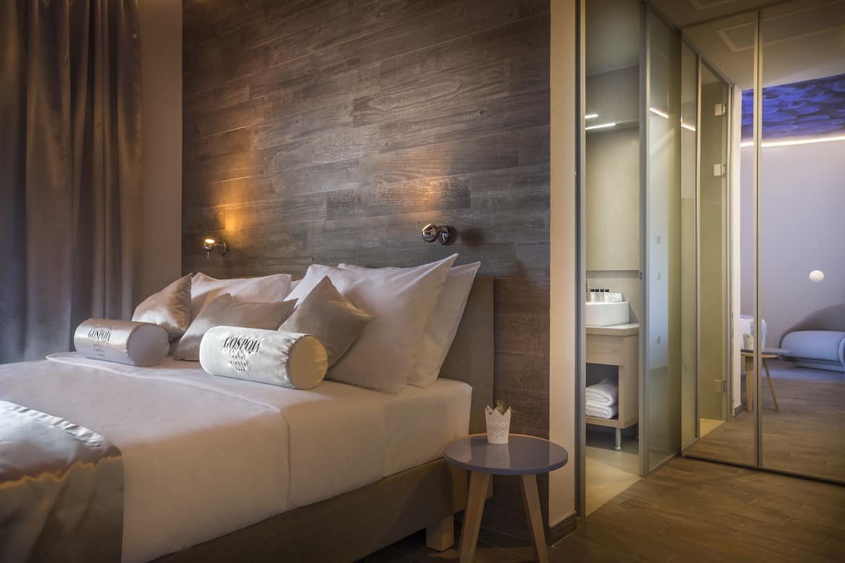 Una habitación de hotel moderna con una cama grande, una mesita redonda pequeña y un separador de vidrio que divide la habitación del baño.