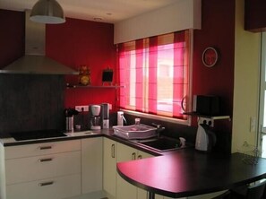 Une cuisine de taille réduite avec des meubles blancs, un plan de travail noir, un mur rouge, une fenêtre ornée de rideaux roses et une horloge au mur.
