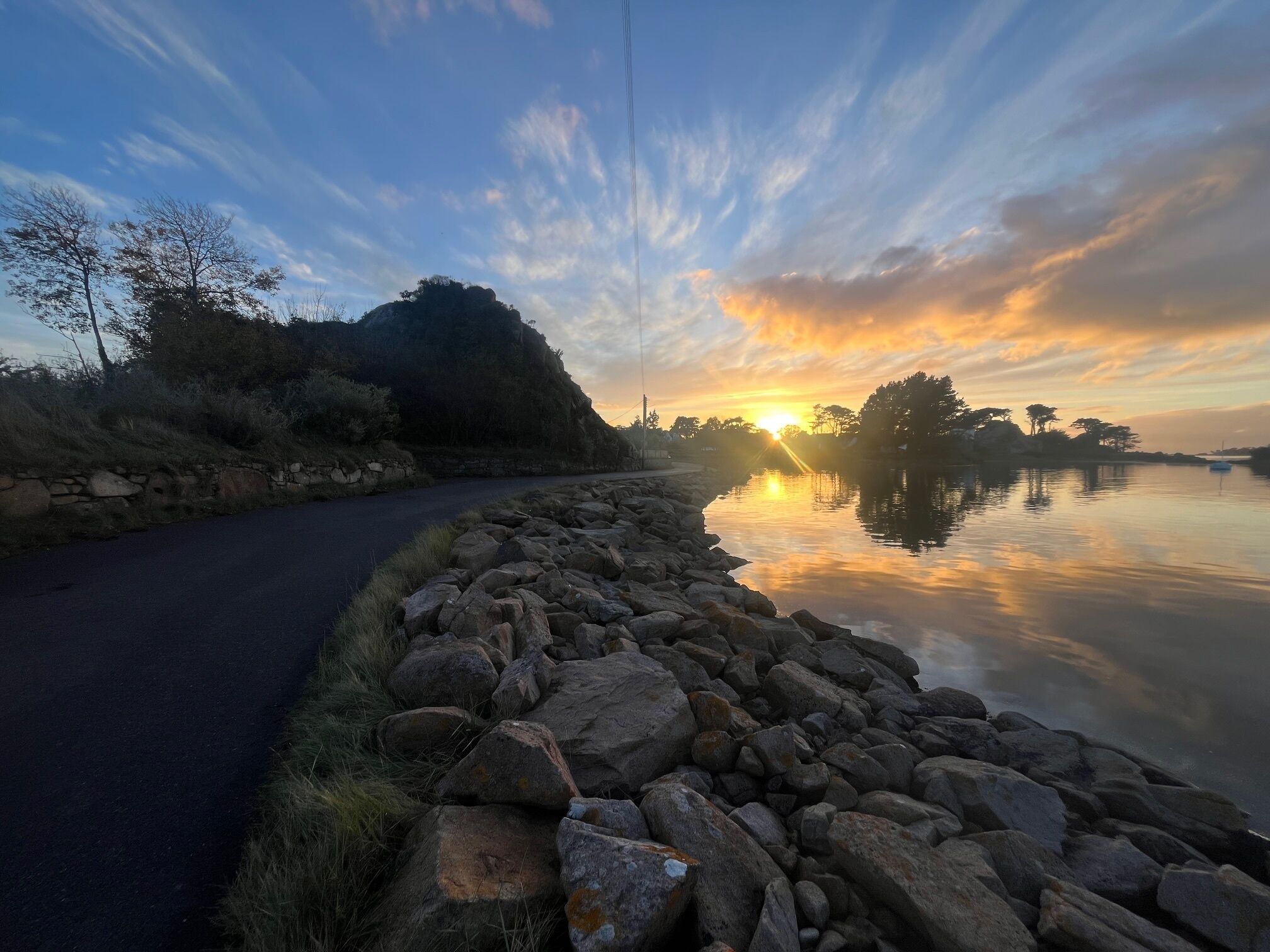 Une route paisible qui longe un lac, avec un mur de pierre d’un côté et une étendue d’eau calme qui reflète le coucher de soleil.