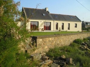 Une maison à deux étages, avec un mur de pierre à l’avant, une cour verdoyante et un ciel dégagé.