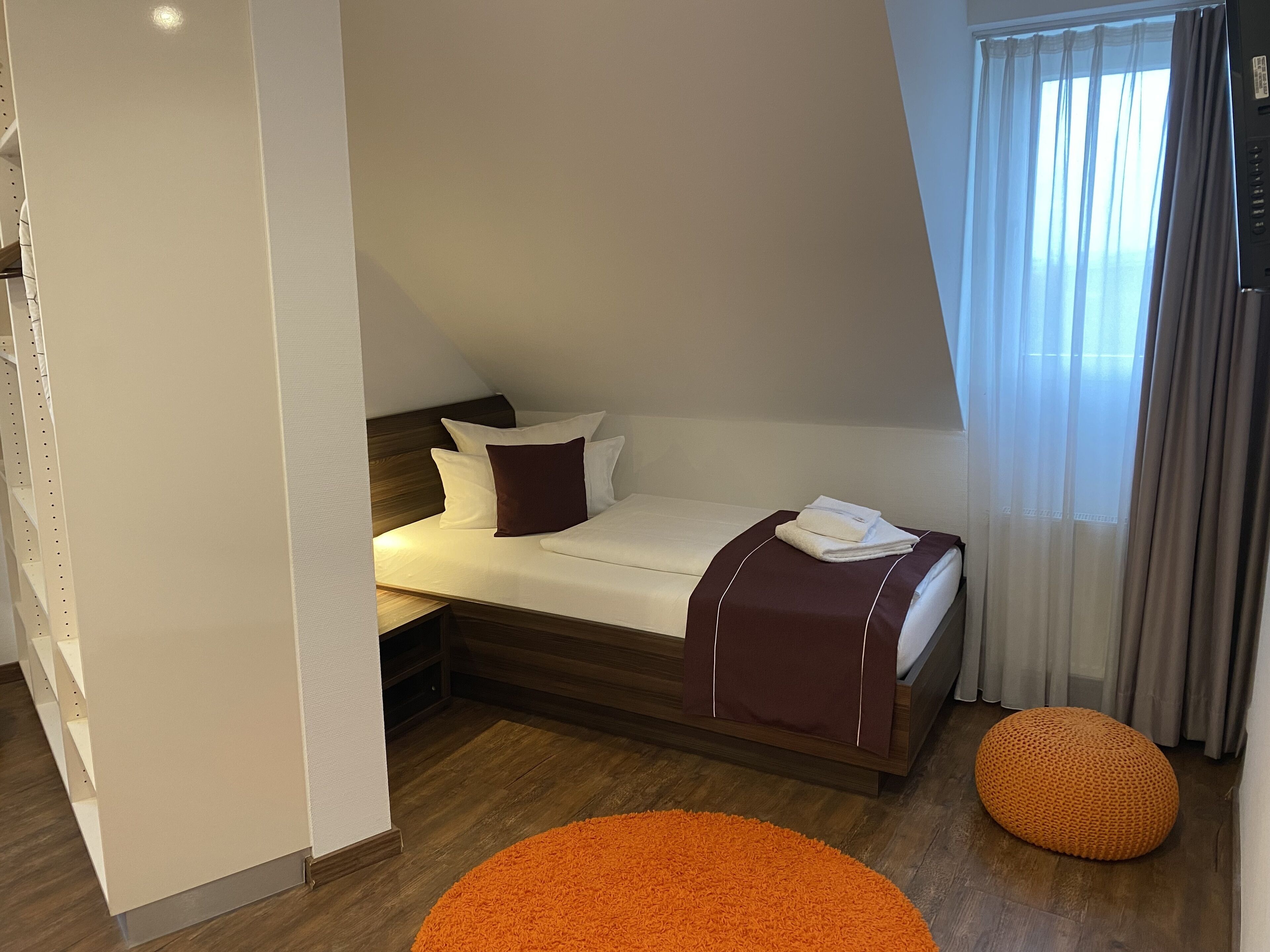 Habitación de hotel moderna con cama, cabecera de madera, pequeño taburete naranja y ventana con cortinas.